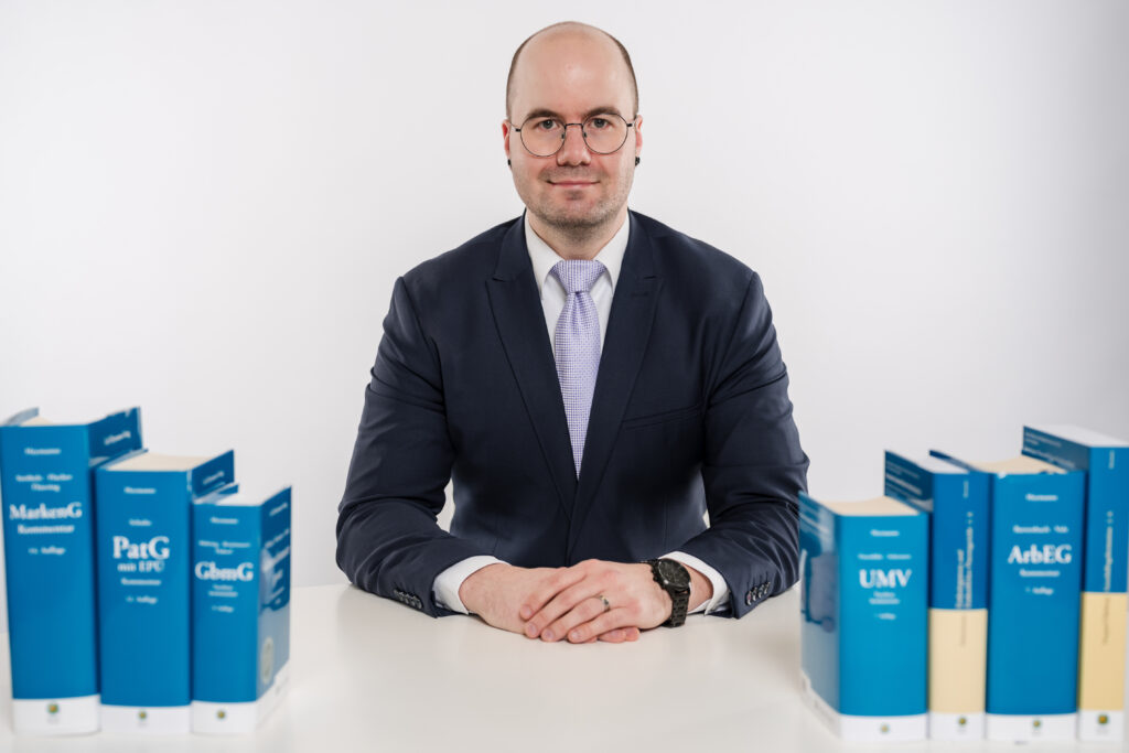 Patentanwalt Philipp Avon sitzt vertrauensvoll an einem Schreibtisch mit Fachbüchern